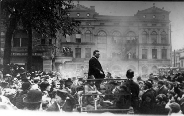 Black and white historical postcard of Teddy Roosevelt giving a speech on the Public Square in Piqua, Ohio. He is on a platform and a large crowd of people are gathered around him.