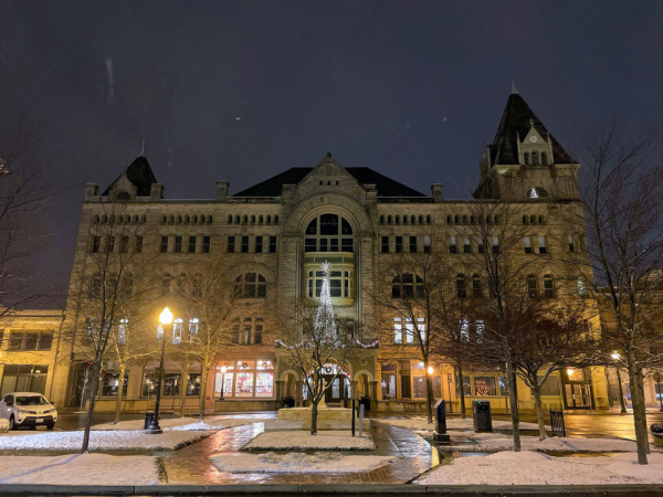A serene photograph of the Fort Piqua Plaza at night. The sky is a deep purple gray color and snow is on the ground and falling. Street lights are lit. There is a tree of lights on the outside of the building, over the front door entrance.