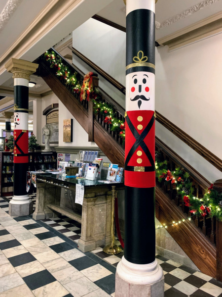 Photograph of a column in the entrance of the Piqua Public Library. The column is decorated to look like a giant toy soldier with a red jacket, gold buttons and a black hat and pants. 