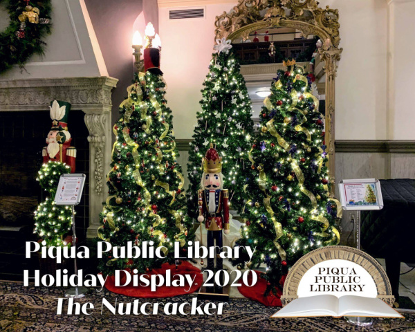 Photograph of three Christmas trees in a group together in the Piqua Public Library lobby. All three trees are decorated with lights and decorations. A large wooden nutcracker stands in front of the middle tree. White text reads "Piqua Public Library Holiday Display 2020 The Nutcracker" and the Piqua Public Library logo is in the bottom right corner. 