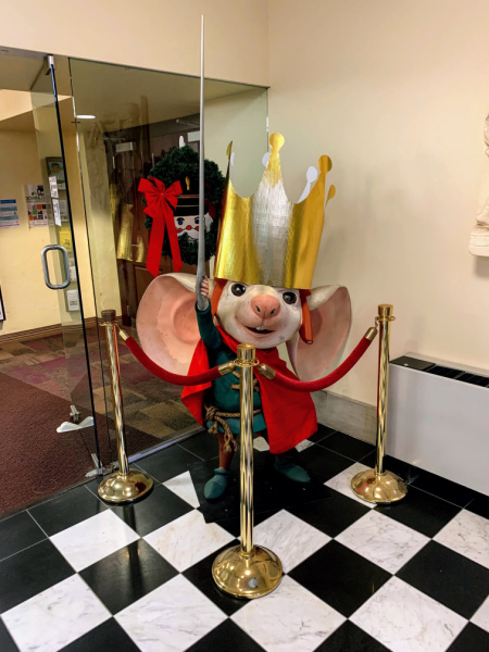 Photograph of a statue of the mouse, Despereaux. He is wearing a red cape and a large gold crown. He holds a sewing needle in the air like a sword.