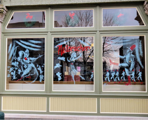 Photograph of the Piqua Public Library front window. The window is painted with white and red paint, illustrating three scenes from The Nutcracker. The left window shows the Rat King with his army of rats. The middle window shows Clara dancing and holding a nutcracker. The Nutcracker is written above her. The right window shows The Nutcracker and his nutcracker army.