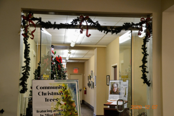 Photograph of the entrance to the Rollin Art Gallery. There is a sign in the foreground which reads Community Christmas Trees. A sign in the background has a photo of library employee and featured artist, Missy Hines. 