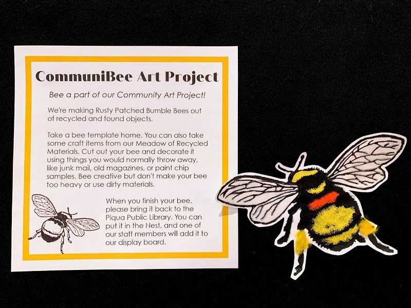 Photograph of a sign with instructions for the CommuniBee Art Project. Next to it is a bumblebee decorated with recycled materials. 