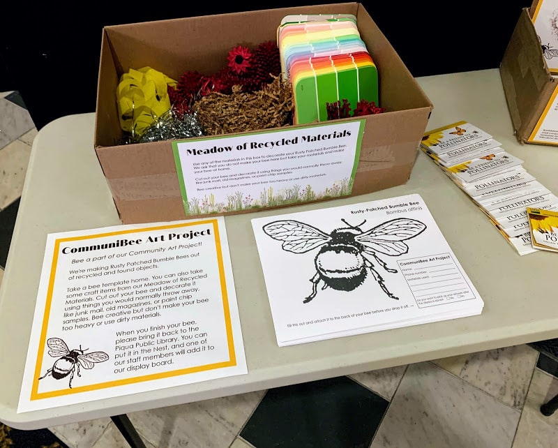Photograph of a table with instructions for the CommuniBee Art Project. Next to it is a bumblebee template and a box of recycled items to be used as craft supplies.
