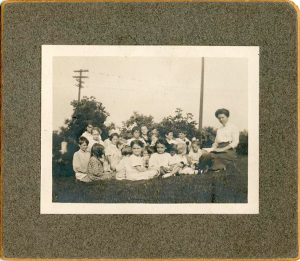 Historical photograph of story time in 1911. A woman in a long dress is seated in the grass next to approximately 20 young children.